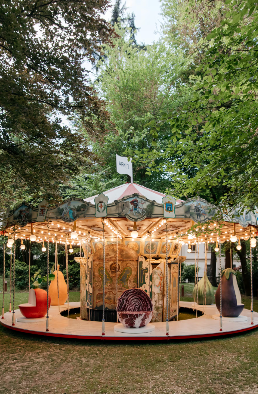 Surreal carousel installation with oversized sculptural fruits and vegetables in a garden setting during Milan Design Week